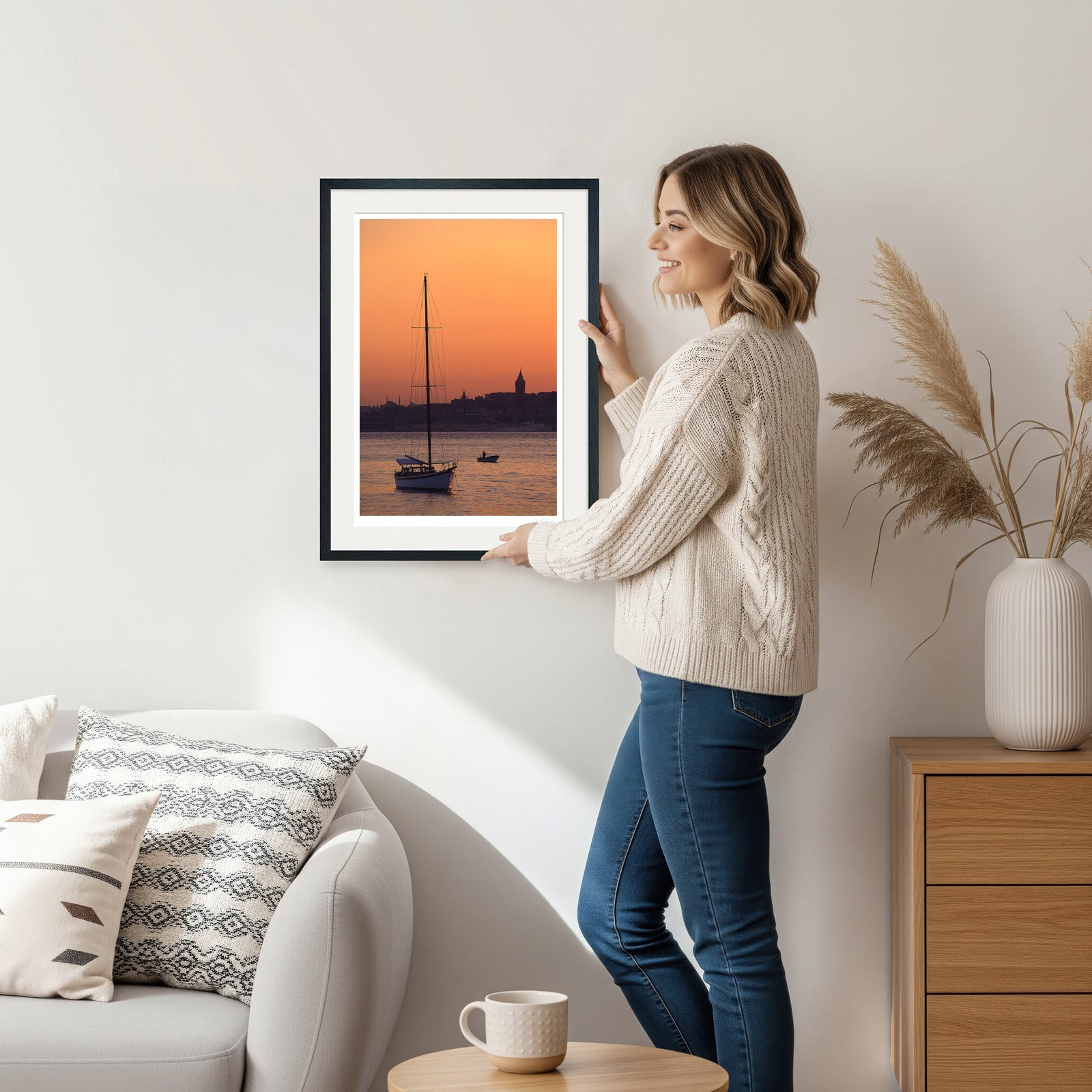 Woman holding black framed sailboat sunset print featuring Galata Tower in her living room setting