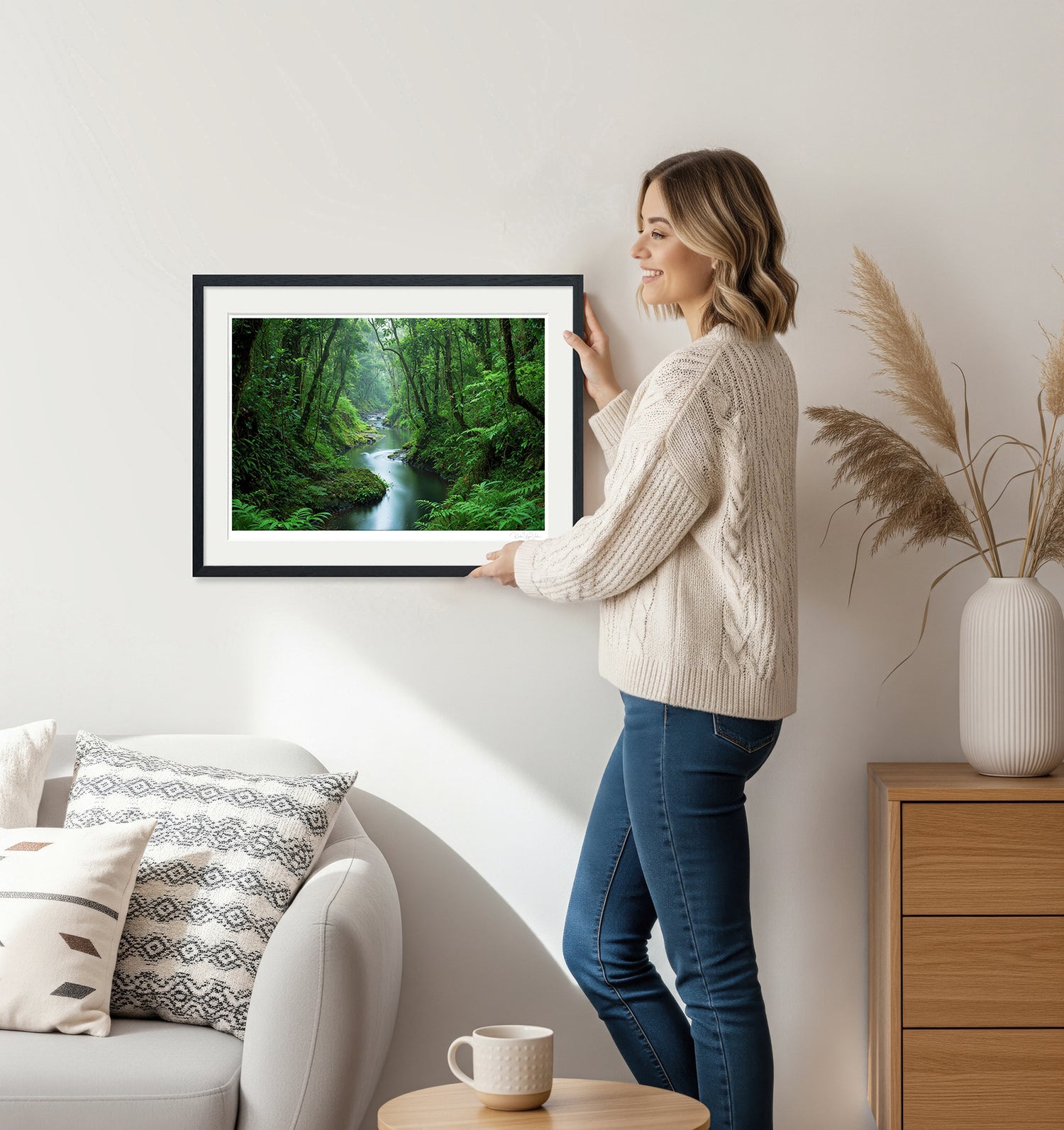 Woman holding framed forest river artwork in black frame – natural decor setting with neutral tones