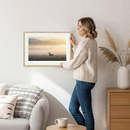 Woman holding a framed deer print with natural oak frame, standing in a cozy living room decorated in neutral tones.