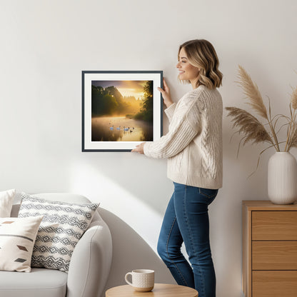 Woman holding framed golden hour lake scene with swans – square format art print in black frame