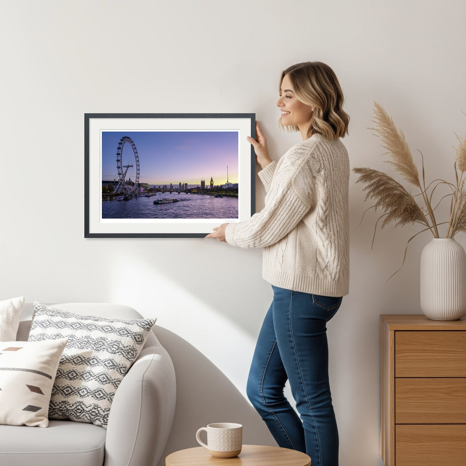 Woman holding a framed twilight London skyline art print featuring the London Eye and Big Ben, standing in a cozy interior with warm lighting.