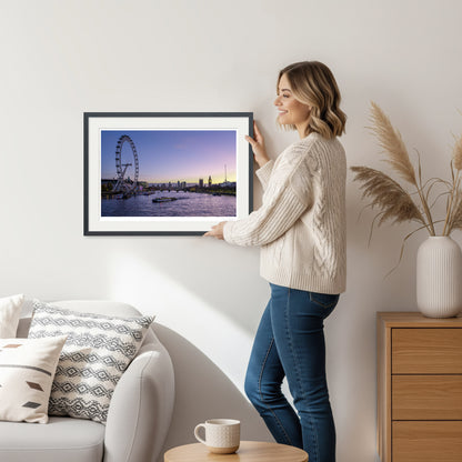 Woman holding a framed twilight London skyline art print featuring the London Eye and Big Ben, standing in a cozy interior with warm lighting.