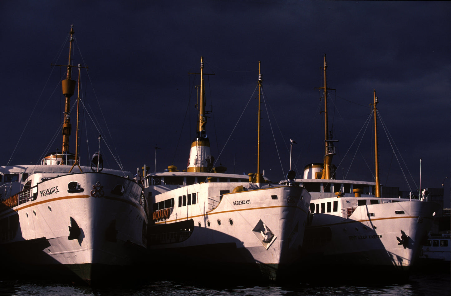 Three classic white and gold Istanbul Bosphorus ferries — Paşabahçe, Sedefadası — moored side by side at harbour against a dramatic dark sky, historic fine art photography print